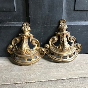 May include: Two antique brass decorative hardware pieces. Each piece has an ornate, symmetrical design with a central vertical element and curved, scroll-like details. The brass has a warm, aged patina, suggesting vintage or reclaimed origin.