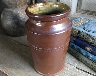 May include: A brown ceramic jar with a wide opening and a slightly flared base. The jar has a textured surface and a decorative band around the middle. The interior has a green glaze. The jar is sitting on a wooden surface.