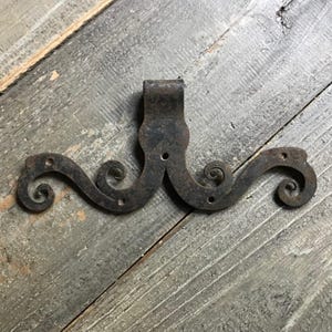 Op de afbeelding: Een donker, verweerd ijzeren beslagstuk met een centrale verticale lip en twee symmetrische, horizontaal uitlopende armen met krulvormige uiteinden. Het metalen oppervlak vertoont sporen van oxidatie en bevat vier kleine montagegaten.
