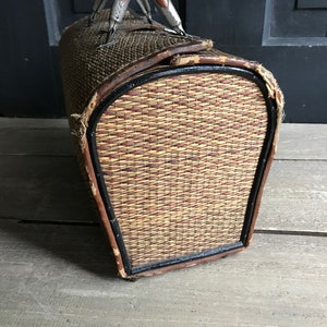 May include: A vintage woven wicker suitcase with a brown leather trim and metal handles. The suitcase has a black painted edge around the opening.