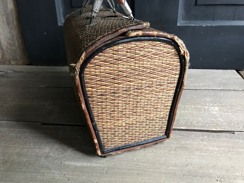 May include: A vintage woven wicker suitcase with a brown leather trim and metal handles. The suitcase has a black painted edge around the opening.