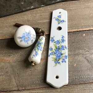 May include: A set of three white porcelain door knobs with a floral design featuring blue forget-me-not flowers. The knobs are in various shapes and sizes, including a round knob, a long knob, and a rectangular backplate.