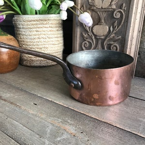 May include: A small copper saucepan with a dark brown handle. The saucepan has a worn, aged look and is sitting on a wooden surface.