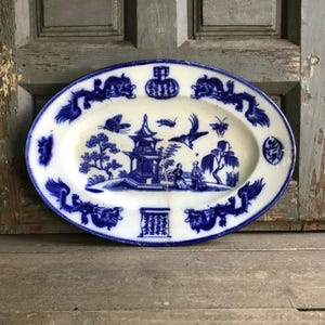 May include: An oval, antique porcelain platter with a blue and white Chinoiserie design. The platter features a central scene with a pagoda, figures, and birds, surrounded by dragons and other decorative elements.