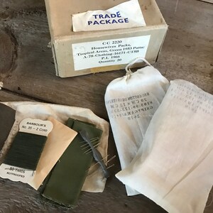 May include: A set of sewing supplies in white cotton bags. The bags contain green thread, a green fabric patch, and needles. The bags are labeled with the text "BARBOUR'S No. 35 - 2 CORD" and "50 YARDS ROTPROOFED". The bags are also labeled with the text "TRADE PACKAGE CC 2220 Housewives Packs, Tropical Areas, Green 1953 Patta A-78-Clothing-16171-CTSH P.L. 1966 Quantity 20".