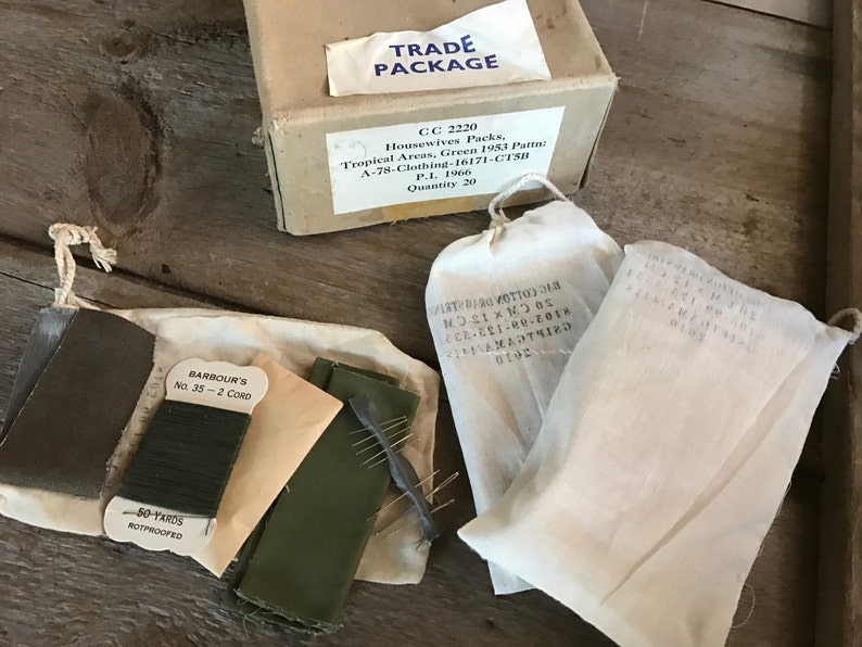May include: A set of sewing supplies in white cotton bags. The bags contain green thread, a green fabric patch, and needles. The bags are labeled with the text "BARBOUR'S No. 35 - 2 CORD" and "50 YARDS ROTPROOFED". The bags are also labeled with the text "TRADE PACKAGE CC 2220 Housewives Packs, Tropical Areas, Green 1953 Patta A-78-Clothing-16171-CTSH P.L. 1966 Quantity 20".