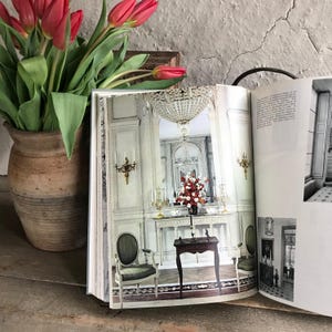 Puede incluir: Un libro abierto muestra una fotografía de una habitación con una lámpara de araña y muebles ornamentados. Un jarrón de tulipanes rojos está a la izquierda. El libro descansa sobre una superficie de madera, con una pared texturizada al fondo. La habitación tiene un arreglo floral.