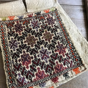 May include: A white fabric pouch with a colorful geometric pattern embroidered on the front. The pouch has a flap with two small tabs.
