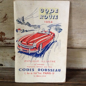 Könnte beinhalten: Ein Vintage-Buchcover für die Ausgabe 1954 des französischen "Code de la Route" (Straßenverkehrsordnung). Das Cover zeigt ein rotes Auto, das auf einer kurvigen Straße fährt, mit einer Berglandschaft im Hintergrund. Der Text "EXPLIQUE - ILLUSTRE" (Erklärt - Illustriert) ist unter dem Auto gedruckt. Das Buch wird von "CODES ROUSSEAU" mit einer Adresse in Paris herausgegeben.