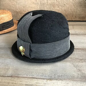 May include: A black straw hat with a wide black and white striped ribbon and a green and gold pin.