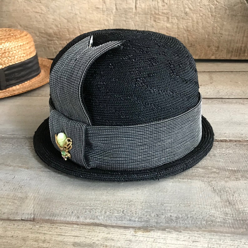 May include: A black straw hat with a wide black and white striped ribbon and a green and gold pin.
