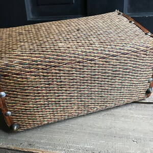 May include: A woven wicker basket with a brown leather trim and silver metal accents. The basket is made of natural materials and has a rustic look.