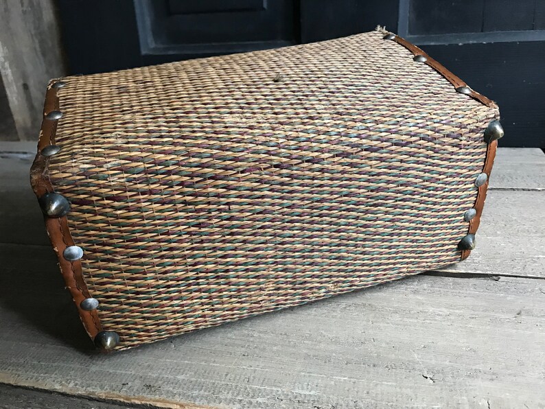 May include: A woven wicker basket with a brown leather trim and silver metal accents. The basket is made of natural materials and has a rustic look.