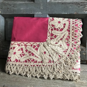 Op de afbeelding: Een gevouwen, rechthoekig textiel met een felroze middenstuk en een crèmekleurige kanten rand. Het kant heeft bloemen- en geometrische patronen, met een geschulpte franje langs de rand. Het textiel is waarschijnlijk een decoratief item.