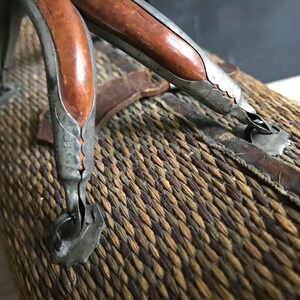 May include: Close-up of a woven wicker suitcase with two leather-wrapped handles. The handles are made of metal with a decorative design and have a brown wooden insert.