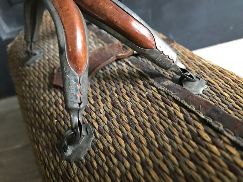 May include: Close-up of a woven wicker suitcase with two leather-wrapped handles. The handles are made of metal with a decorative design and have a brown wooden insert.