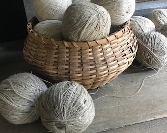 May include: A woven basket filled with several balls of yarn. The yarn balls are various shades of gray and beige. The basket is made of light brown woven material and sits on a weathered wooden surface.