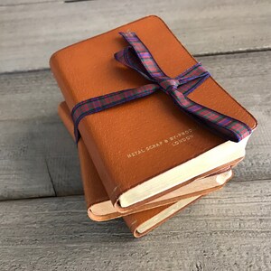 May include: Three brown leather bound journals tied together with a red, blue, and green plaid ribbon. The top journal has the text "METAL SCRAP & BY-PRODUCT LONDON" embossed on the front cover.
