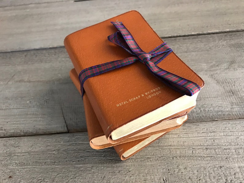 May include: Three brown leather bound journals tied together with a red, blue, and green plaid ribbon. The top journal has the text "METAL SCRAP & BY-PRODUCT LONDON" embossed on the front cover.