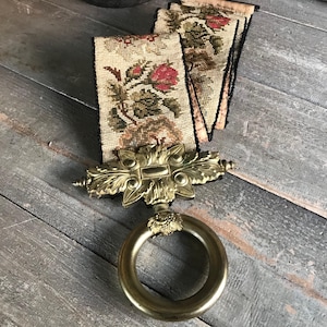 Puede incluir: Un recogedor de cortina antiguo de latón con diseño floral. El recogedor presenta un anillo decorativo y un adorno floral detallado. Se adjuntan dos paneles de tela bordada con rosas rojas y hojas verdes.