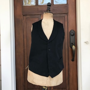 May include: A black sleeveless vest with a shawl collar and five buttons. The vest is on a mannequin in front of a wooden door.