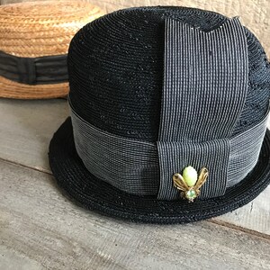 May include: A black straw hat with a wide brim and a black and white striped ribbon. The hat has a small green and gold brooch in the shape of a bug.