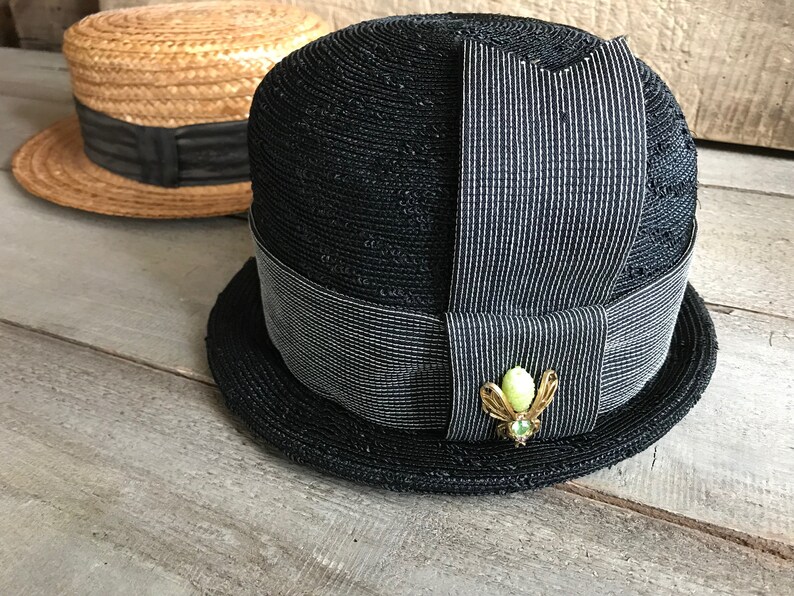 May include: A black straw hat with a wide brim and a black and white striped ribbon. The hat has a small green and gold brooch in the shape of a bug.