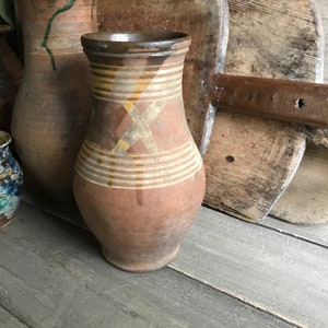 May include: A brown clay jug with yellow and brown painted stripes and a cross pattern. The jug is sitting on a wooden surface.
