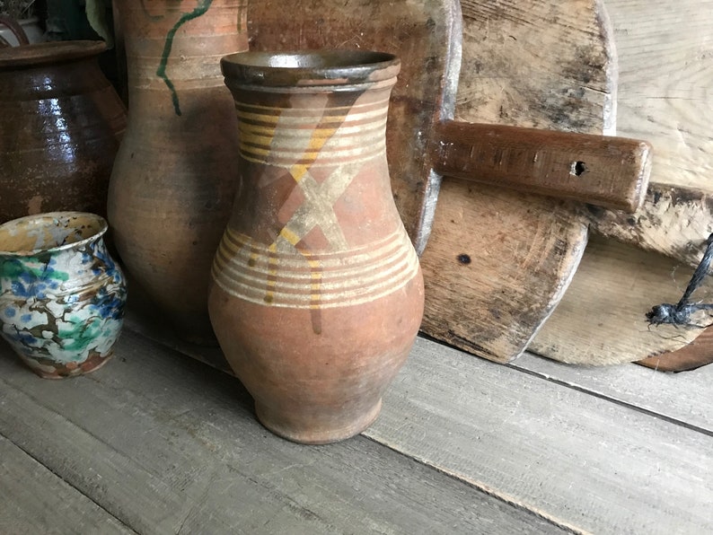 May include: A brown clay jug with yellow and brown painted stripes and a cross pattern. The jug is sitting on a wooden surface.