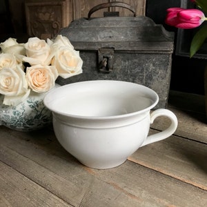 May include: A white ceramic chamber pot with a handle. The pot is sitting on a wooden surface.