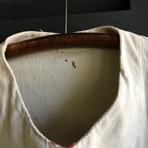 K&ouml;nnte beinhalten: Nahaufnahme eines cremefarbenen Kleidungsst&uuml;cks mit V-Ausschnitt, das an einem Holzb&uuml;gel h&auml;ngt. Der Stoff hat ein strukturiertes Aussehen. Auf dem Kleidungsst&uuml;ck befinden sich kleine braune Flecken. Der Kragen ist dunkler.
