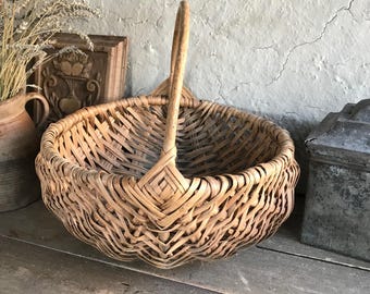 May include: A large, round, woven basket with a single handle. The basket is made of natural brown wicker and has a slightly scalloped edge.