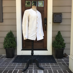 K&ouml;nnte beinhalten: Cremefarbene Jacke mit rundem Kragen und Knopfverschluss, pr&auml;sentiert auf einer schwarzen Schaufensterpuppe. Die Jacke hat lange &Auml;rmel und zwei Vordertaschen. Die Schaufensterpuppe steht auf einem schwarzen St&auml;nder. Der Hintergrund umfasst eine braune T&uuml;r, gr&uuml;ne Topfpflanzen und einen Ziegelweg.
