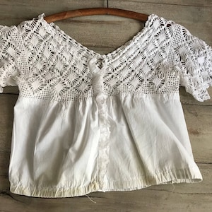 May include: A white, vintage-style blouse with a V-neck and short sleeves. The top features a detailed, openwork crochet yoke and sleeves, with a gathered, plain white cotton body. The blouse is hanging on a wooden hanger.