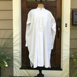 May include: A white long-sleeved shirt with a mandarin collar and a pleated front panel. The shirt is on a mannequin in front of a wooden door.
