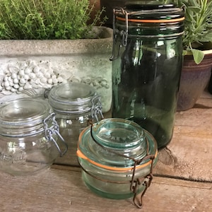 May include: Four glass jars with metal lids and rubber seals. The jars are various sizes and colors, including clear, green, and blue. The jars are sitting on a wooden surface.