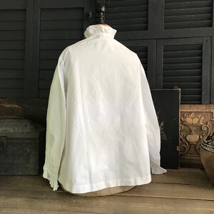 Pu&ograve; includere: Una camicia bianca a maniche lunghe con colletto e polsini a balze. La camicia &egrave; esposta su un manichino sullo sfondo di un'imposta in legno e di un arredamento antico. Il tessuto sembra essere cotone o un materiale simile.