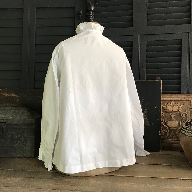 Pu&ograve; includere: Una camicia bianca a maniche lunghe con colletto e polsini a balze. La camicia &egrave; esposta su un manichino sullo sfondo di un'imposta in legno e di un arredamento antico. Il tessuto sembra essere cotone o un materiale simile.