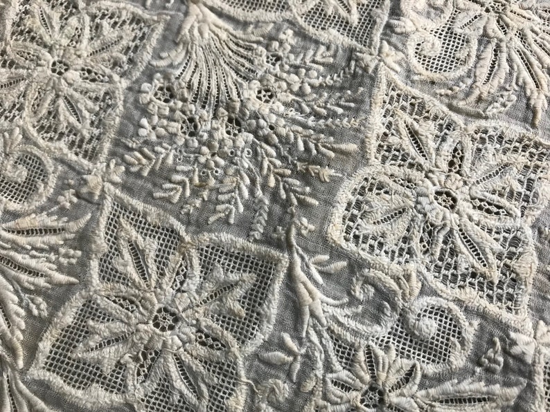 以下が含まれることがあります： 繊細なレースワークと花柄のモチーフが特徴の、複雑な刺繍が施された白い綿生地。