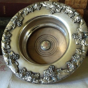 May include: Silver-plated bottle coaster with a grapevine design. The coaster has a wooden center with a circular design.