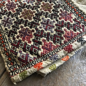May include: A beige woven fabric with a geometric pattern in shades of red, orange, purple, green, and black. The fabric has a fringed edge.