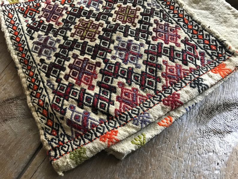 May include: A beige woven fabric with a geometric pattern in shades of red, orange, purple, green, and black. The fabric has a fringed edge.