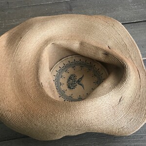 K&ouml;nnte beinhalten: Ein breitkrempiger Strohhut mit brauner gewebter Textur. Die Innenseite des Hutes hat ein gedrucktes Design mit einer Blume und einem Vogel.
