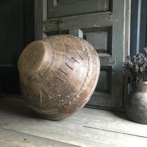 May include: A large, antique-style, brown ceramic vessel with a rounded body and a flat top. The vessel features decorative stitching and is placed on a weathered wooden surface. A smaller vase with lavender is to the right.