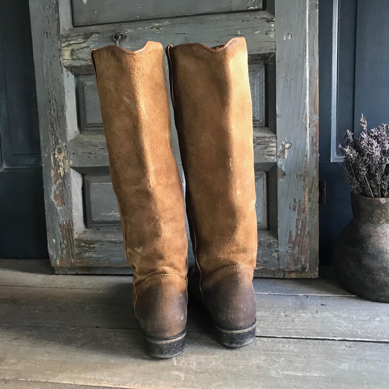 May include: A pair of tall, brown suede boots. The boots have a worn, vintage look with a darker brown color on the soles and lower portion. The boots are standing upright against a weathered wooden door.