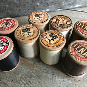 Könnte beinhalten: Eine Sammlung von acht Garnrollen, jede mit einer anderen Farbe und einem anderen Etikett. Die Spulen sind aus Holz und haben verschiedene Etiketten, darunter "Cable Lin", "Cousin Frères", "Tenax" und "Droulers Vernier".