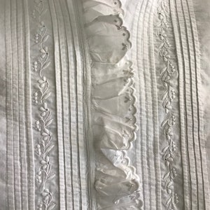 Pu&ograve; includere: Primo piano di un capo bianco con ricami e pieghe dettagliate. Il design presenta pieghe verticali, ricami floreali e un bordo a balze. Il tessuto sembra essere di cotone o lino leggero.