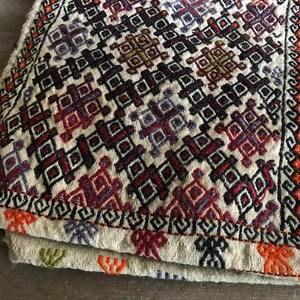 May include: A white woven rug with a geometric pattern in shades of red, orange, black, and purple. The rug has a fringe border.