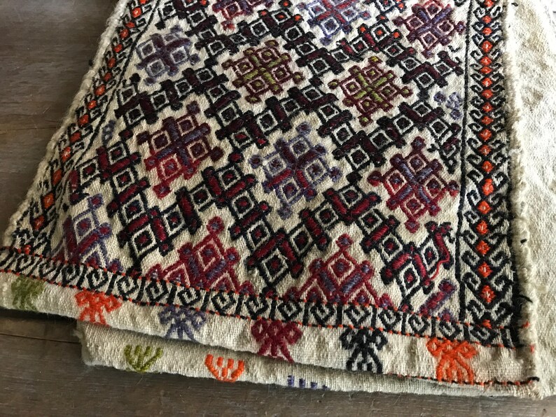 May include: A white woven rug with a geometric pattern in shades of red, orange, black, and purple. The rug has a fringe border.