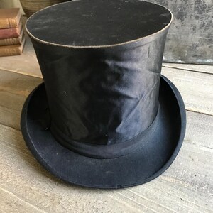 May include: A black top hat with a tall, cylindrical crown and a wide brim. The hat is made of a smooth, black fabric.